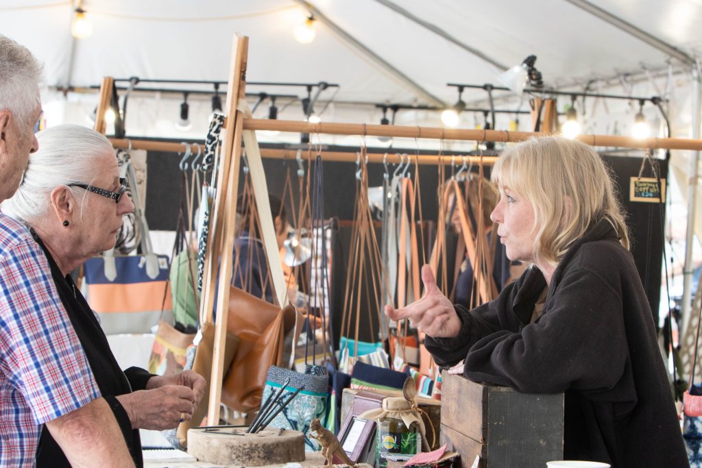 Leather bag artesian gestures as she speaks to a customer on the other side of her booth at the Maker Marketplace.