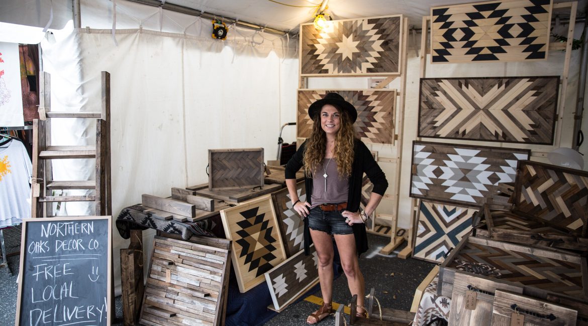 Northern Oaks Decor Company artisan stands in her Maker Marketplace tent with elaborately patterned oak panels.