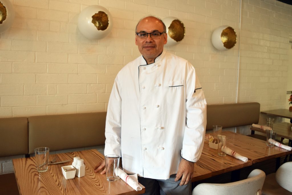 Jorge Castillo standing between the wooden tables of embur fire fusion wearing a white chefs coat and glasses.