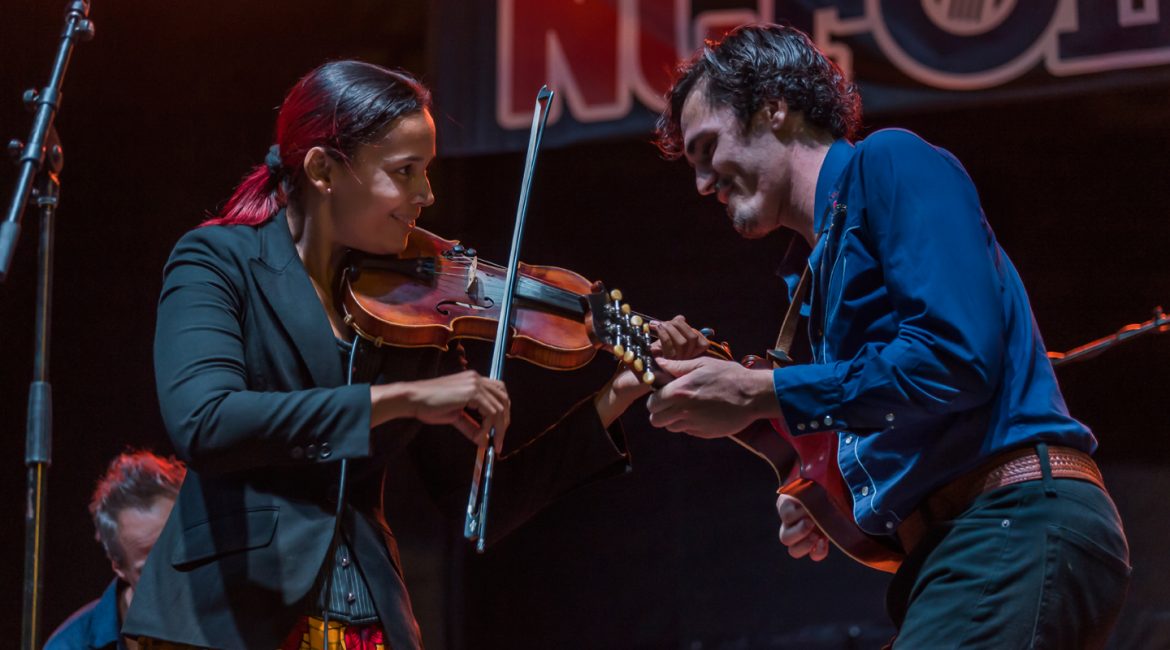 Rhiannon Giddens plays the fiddle as she smiles and leans towards to her fellow musician playing the guitar on a North Carolina Folk Festival stage.