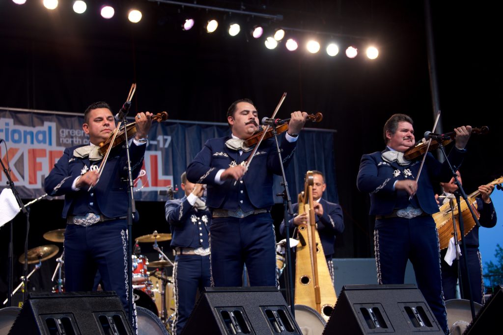 Three members of Mariachi Los Camperos preform on a festival stage in traditional mariachi attire.