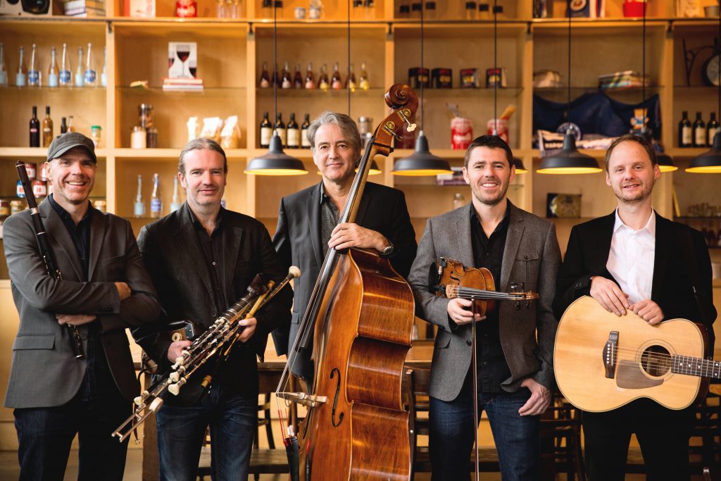 Lúnasa members with their instruments (a guitar, bass, bodhran, fiddle, and uilleann pipes) stand in front of a bar.