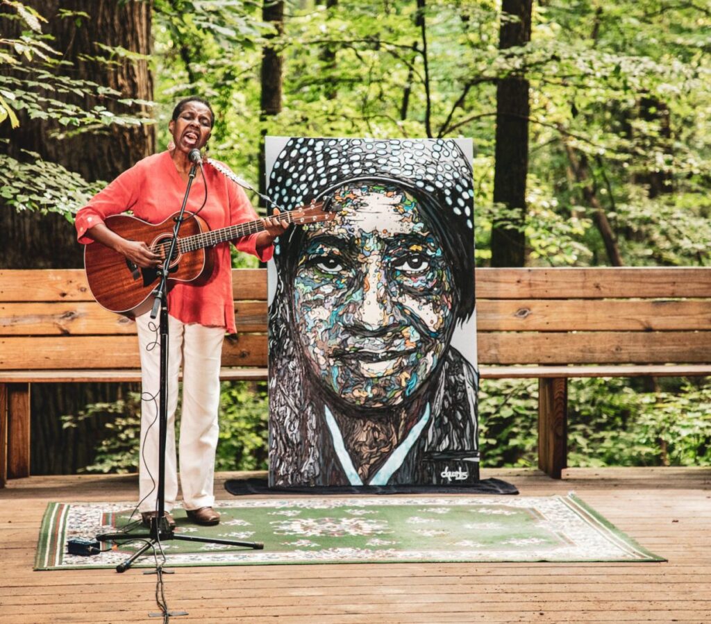 Jackson plays guitar on a deck in the forest. Behind her is a portrait of an African American woman.