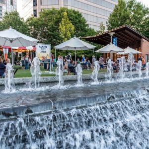 Water fountain in Greensboro's Center City Park in foreground with tents and sign indicating location of Center City Jams in the background. 2022 N.C. Folk Fest