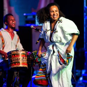 Ethiopian singer dancing on stage in white dress in foreground. Her bandmate and percussionist in background. Performing at 2022 N.C. Folk Fest.